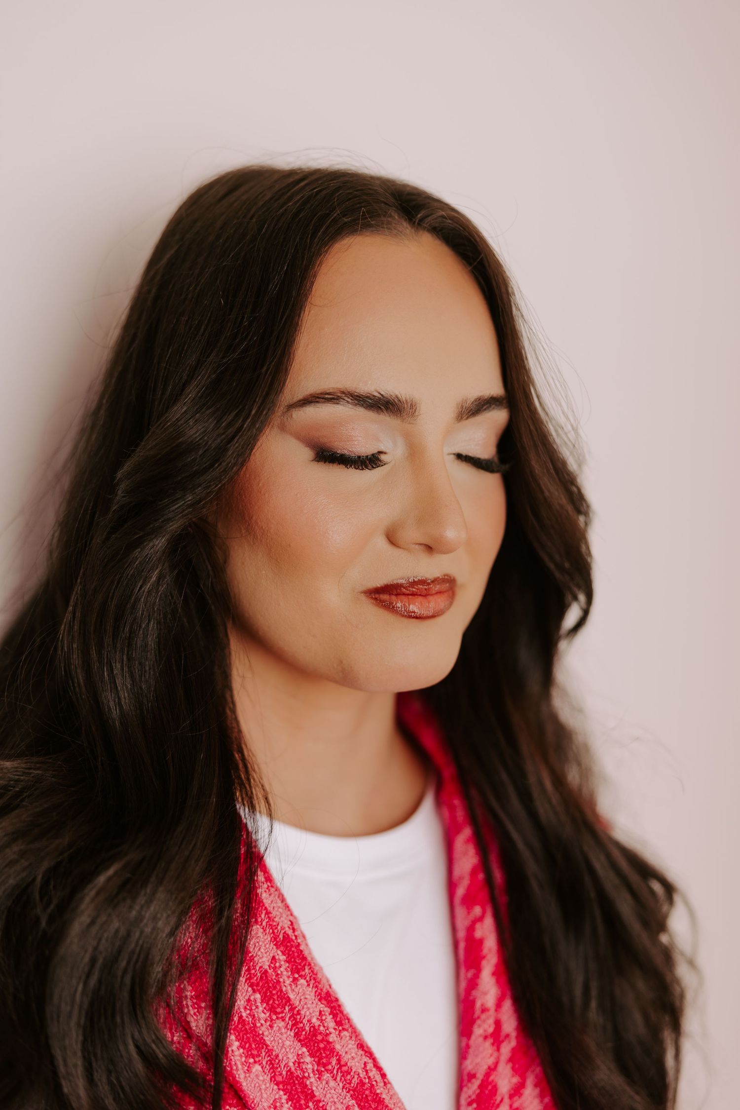 Woman with long dark hair, red lips, and a pink houndstooth scarf poses with eyes closed against a white background.