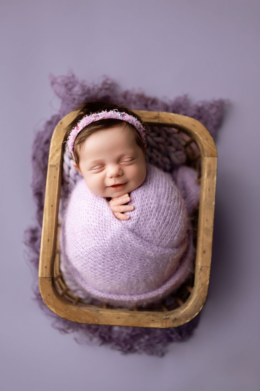 Smiling newborn baby girl swaddled in lavender wrap with floral headband resting in wooden basket.