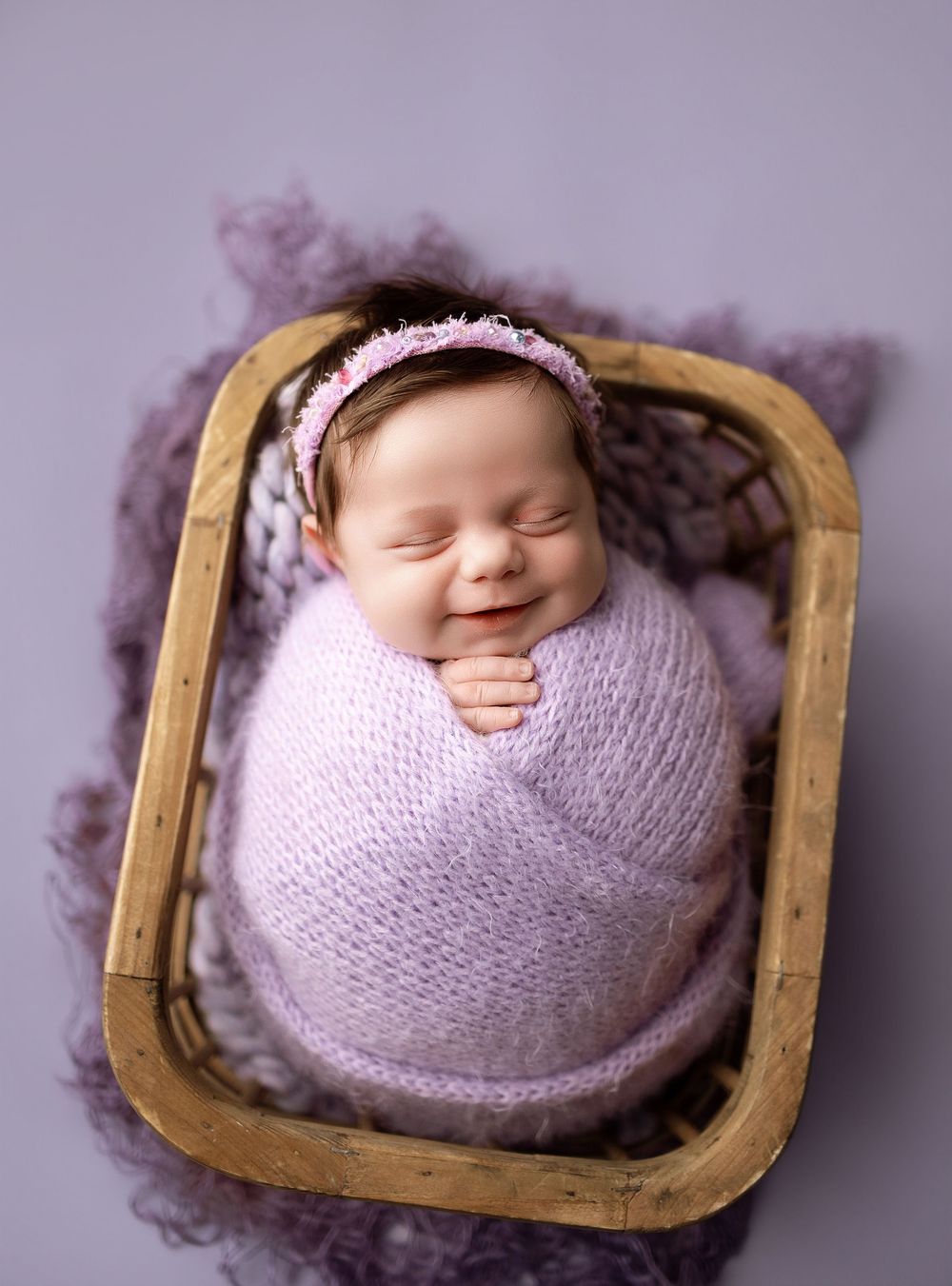 Smiling newborn baby girl swaddled in lavender wrap with floral headband, resting in a wooden basket on purple background.