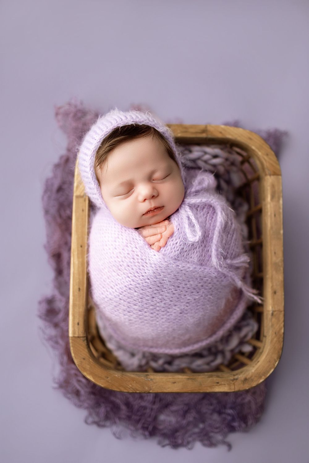Sleeping newborn baby swaddled in purple knit wrap and bonnet, resting in a wooden basket on lavender background.