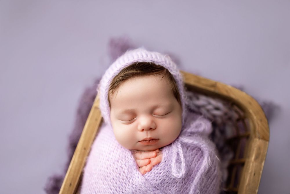 Sleeping newborn baby wrapped in lavender knit swaddle wearing matching bonnet, nestled in wooden basket.