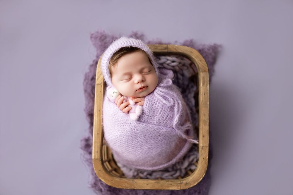 Sleeping newborn baby swaddled in lavender wrap wearing knit bonnet, resting in wooden basket on purple fur.