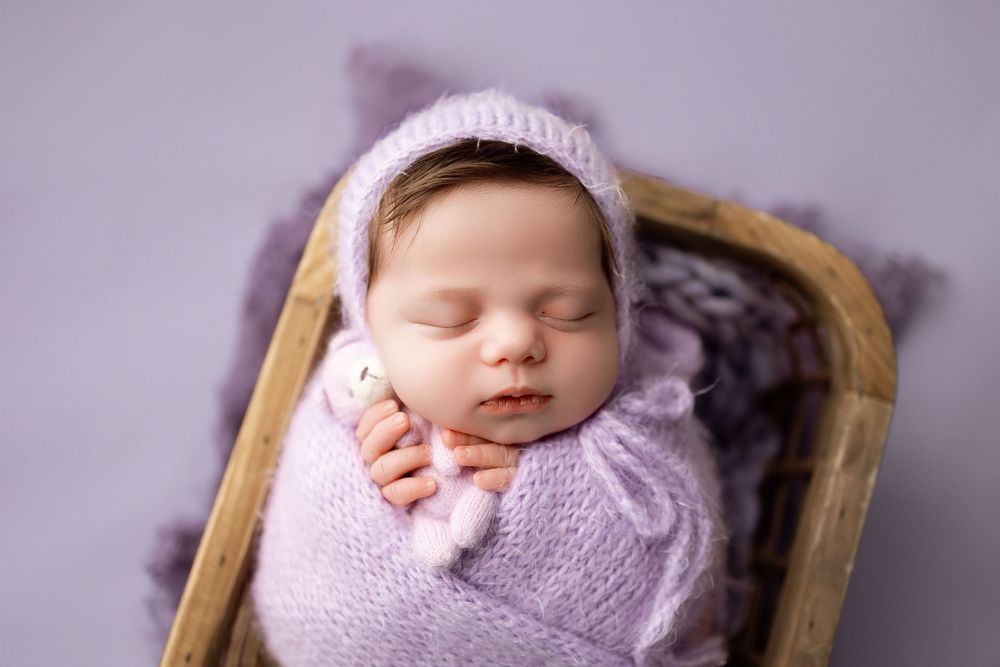 Sleeping newborn baby wrapped in purple knit blanket with matching bonnet in wooden basket.