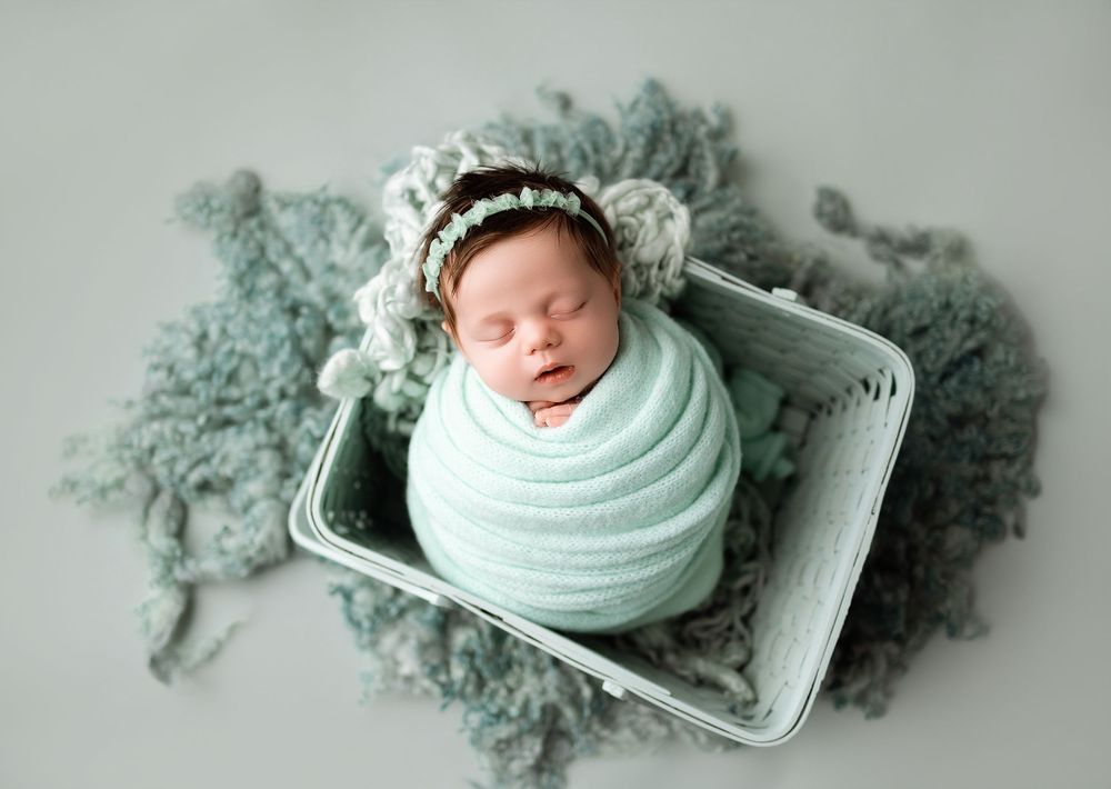 Newborn baby swaddled in mint green wrap, sleeping peacefully in a small box with a lace bonnet on a blue backdrop.