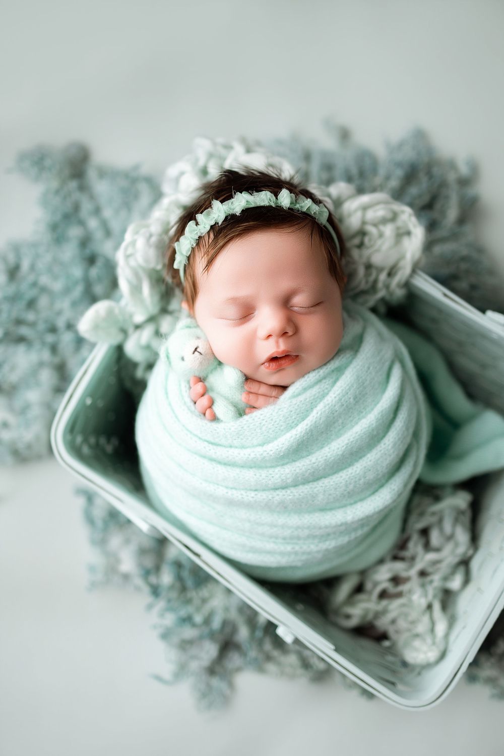 Newborn baby swaddled in mint green wrap, wearing floral headband, sleeping peacefully in a small wooden crate.