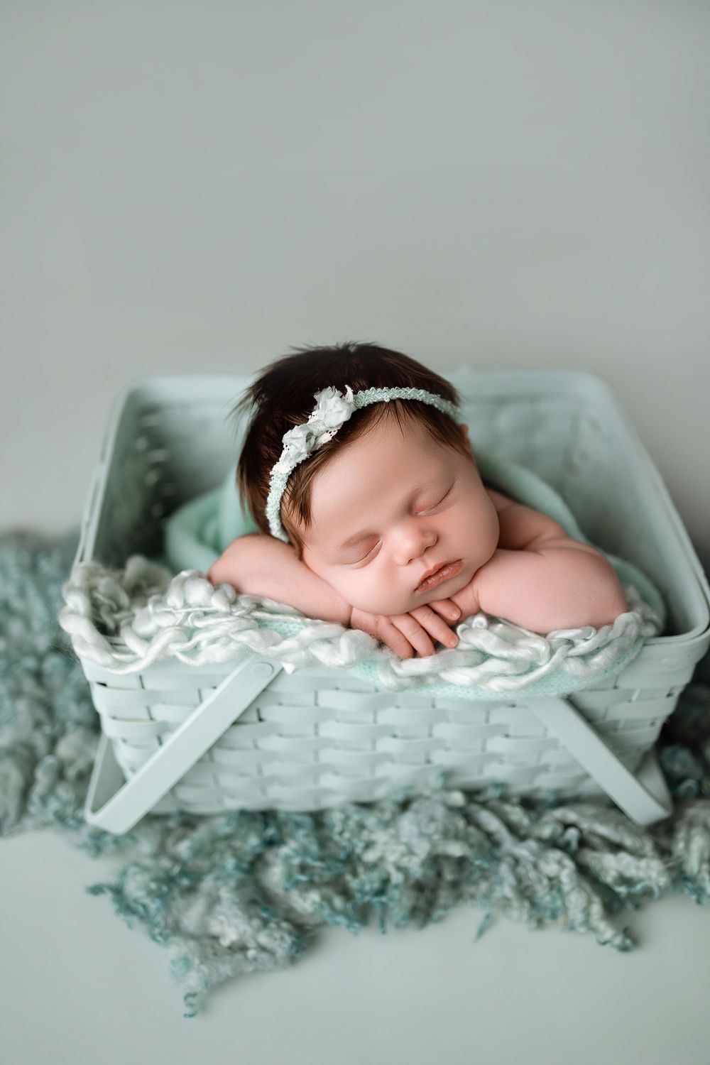 Sleeping newborn baby girl in mint green basket with floral headband, wrapped in soft teal blankets.