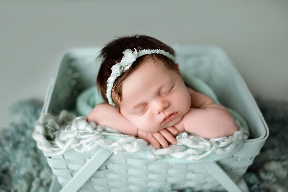 Sleeping newborn baby girl in mint green basket with floral headband on soft teal fur blanket.