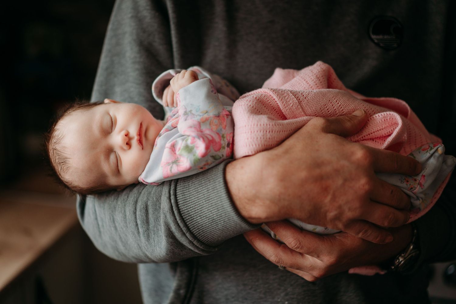 A newborn baby sleeping peacefully while being cradled in adult arms, wrapped in a pink floral blanket.