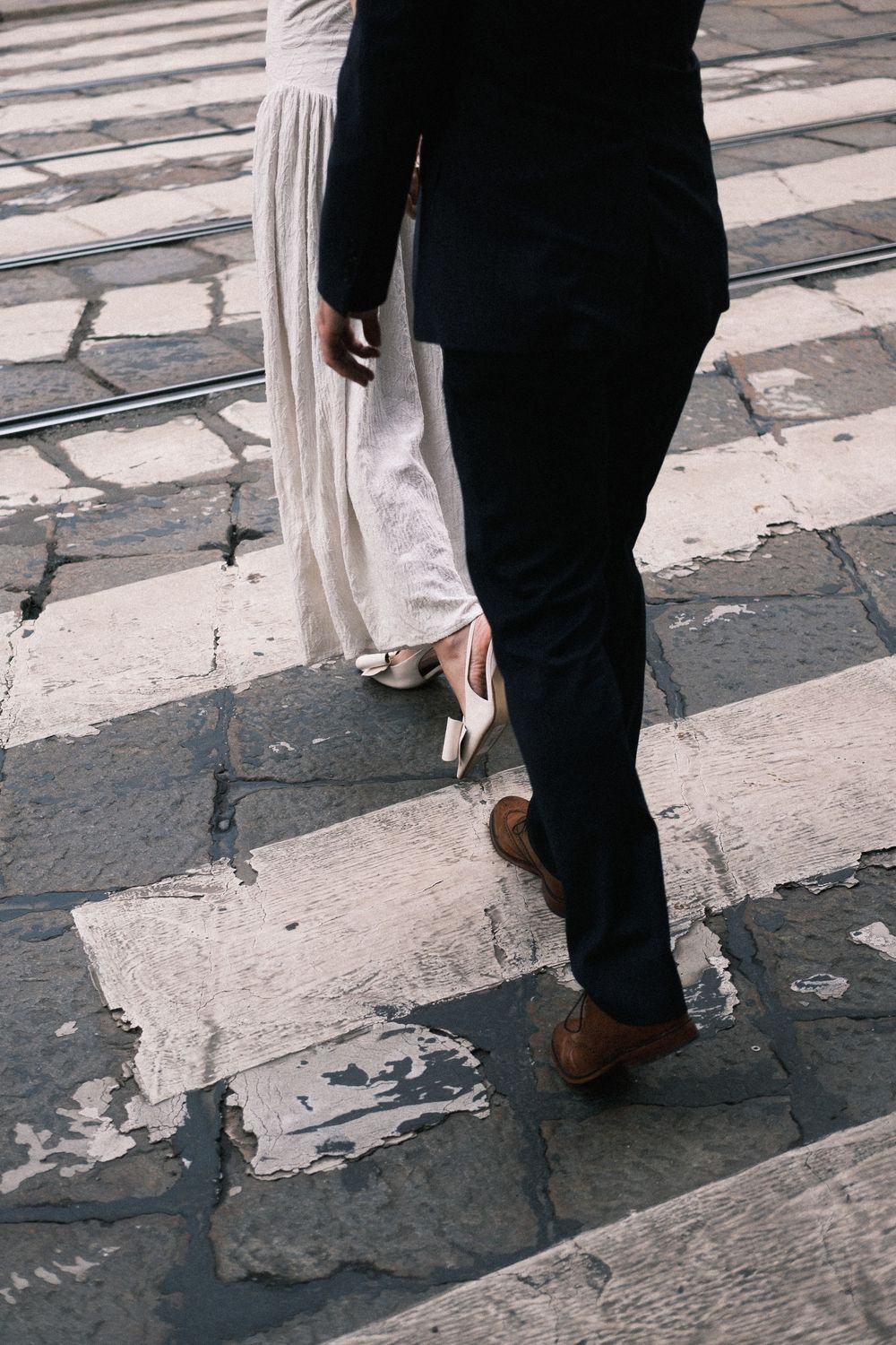Bride in white dress and groom in dark suit walking across cobblestone street, captured from waist down.