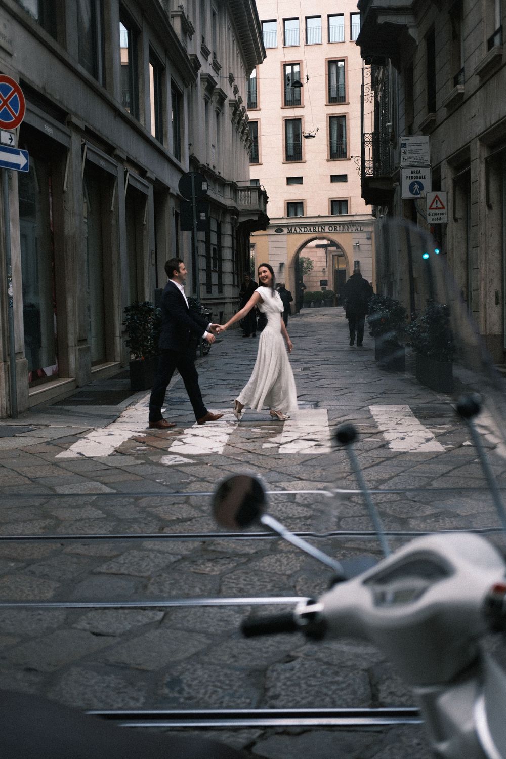 Couple in wedding attire holding hands while crossing a cobblestone street in a European city.