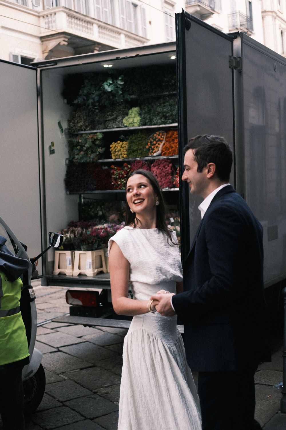 Couple in wedding attire holding hands and smiling in front of a colorful flower delivery truck.