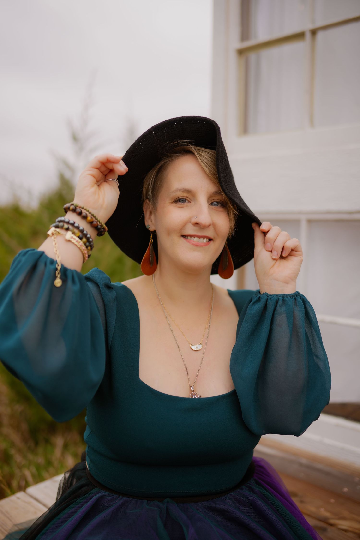 Woman in teal blouse and black hat poses outdoors near a white building.