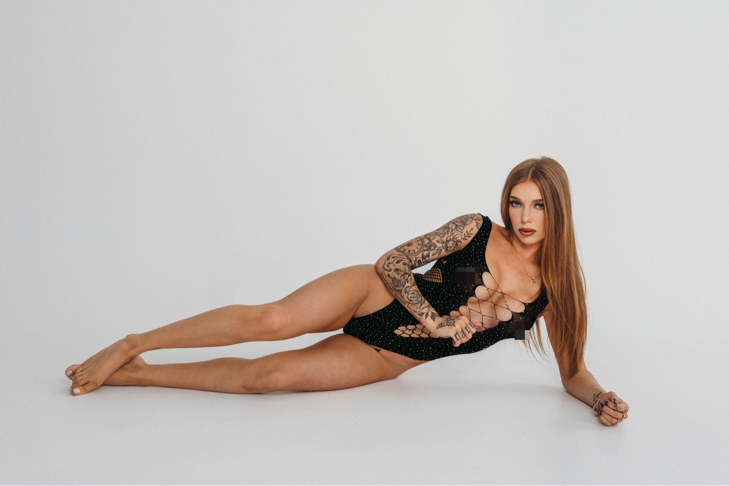 Tattooed woman in black bodysuit posing on white studio floor, showcasing flexibility and body art.