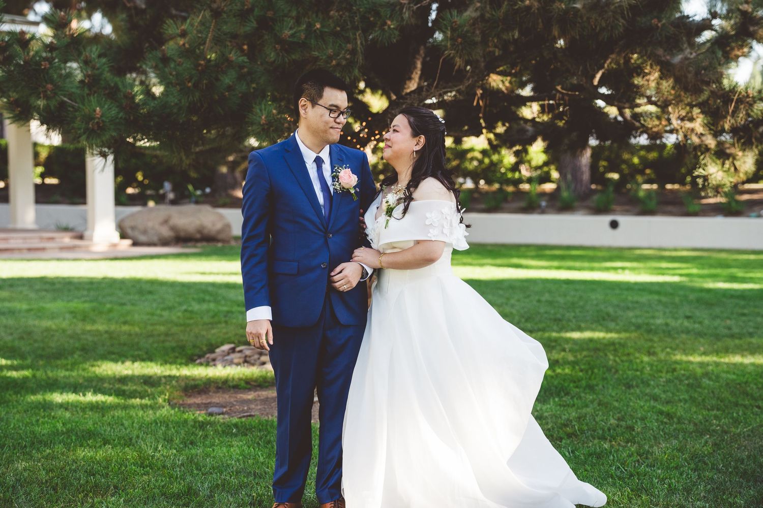 A couple in wedding attire shares a romantic moment on a green lawn, groom in blue suit, bride in white gown.