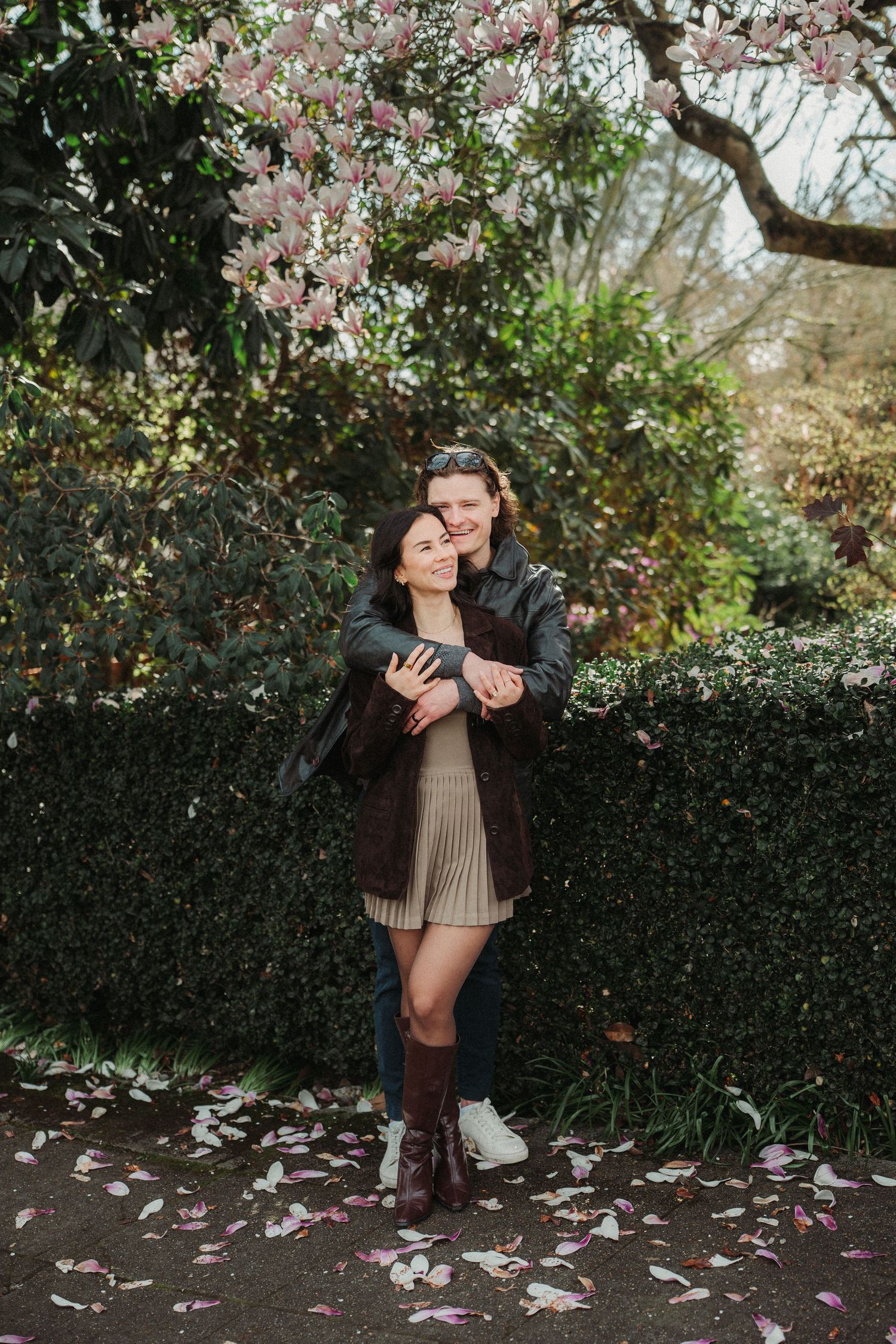 Couple embracing under blooming magnolia tree in garden, woman in beige dress and boots, man in dark jacket.