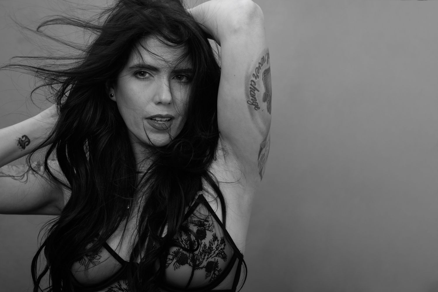 Black and white photo of a tattooed woman with dark hair posing in lace top.