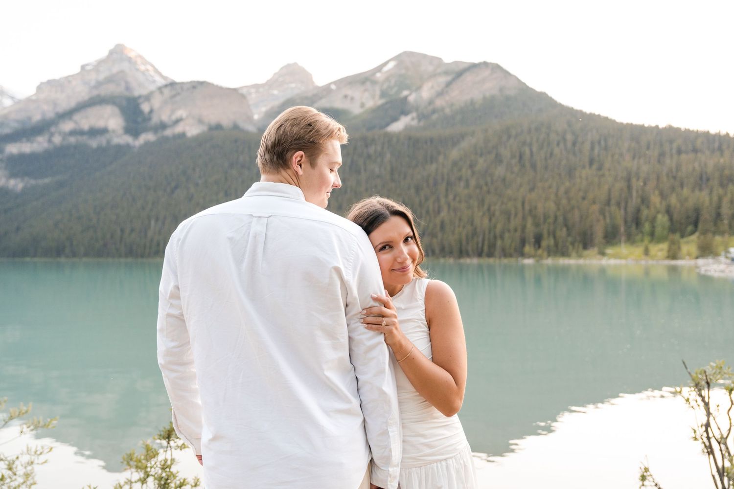 Couple in white attire sharing a romantic moment by a turquoise Lake Louise in Banff, Canada.