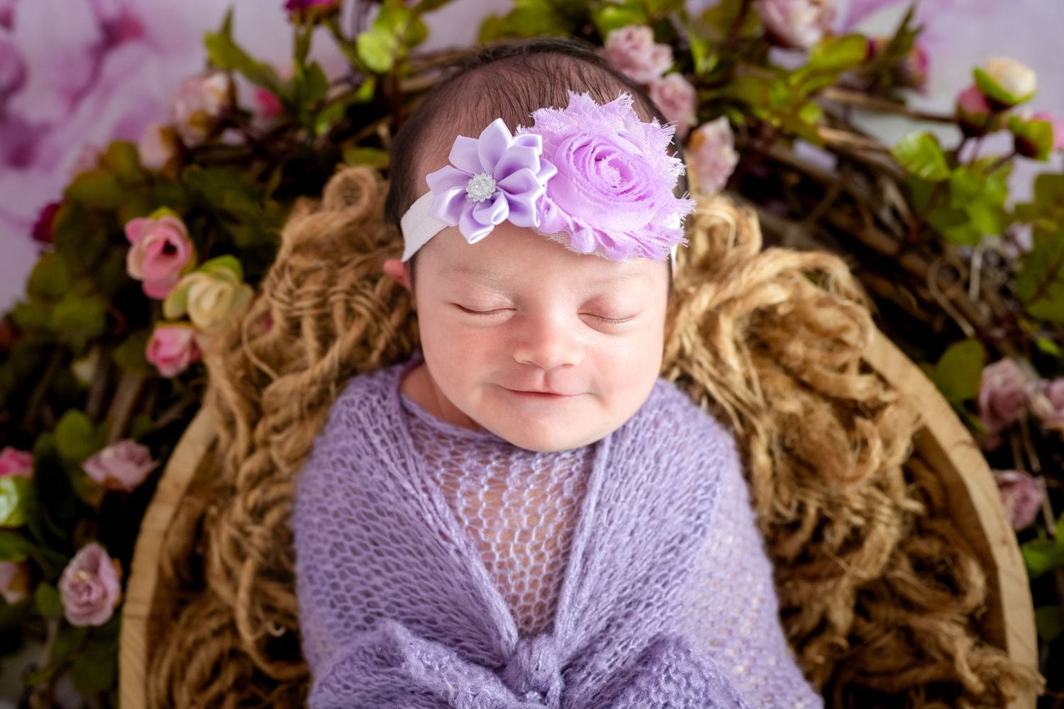 Sleeping newborn baby wrapped in purple knit swaddle with floral headband in a basket surrounded by pink roses.