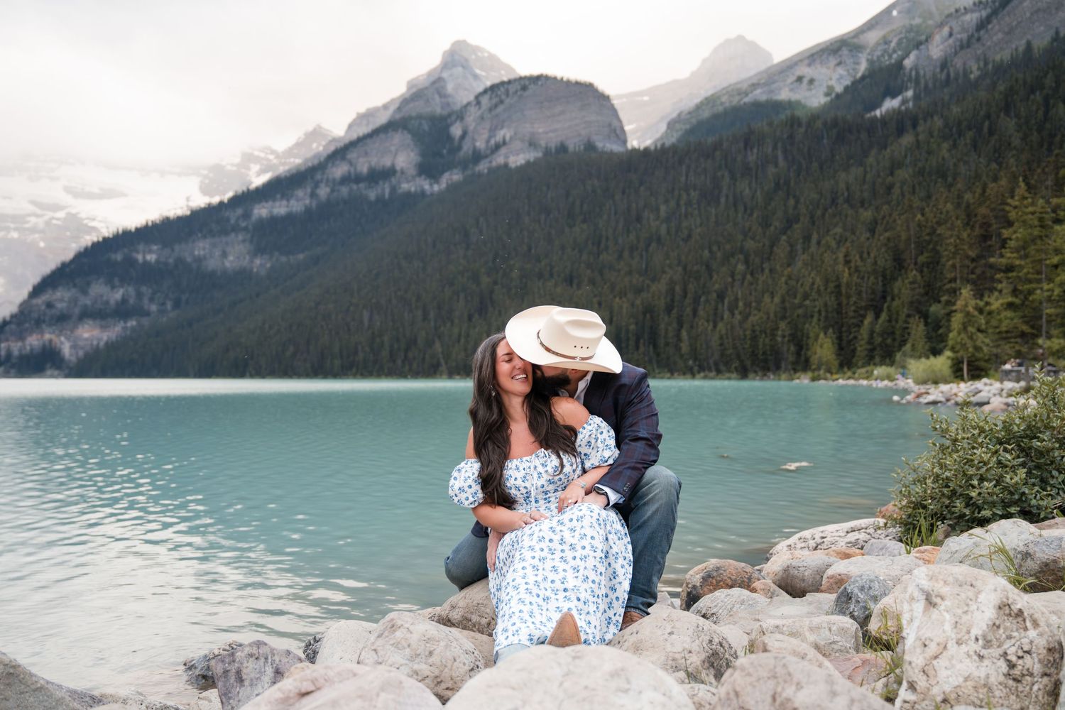 Couple sharing a romantic kiss on rocky shore of turquoise Lake Louise, Banff with mountains and pine trees backdrop.