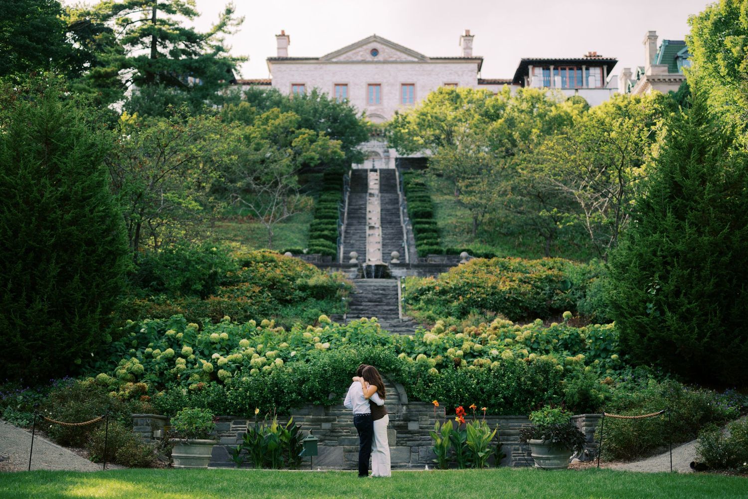 Couple embracing in lush garden with grand staircase and elegant mansion backdrop at a romantic outdoor wedding venue.