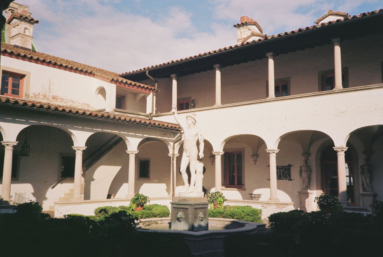 Renaissance courtyard with arched colonnades, central fountain sculpture, and lush greenery in a historic Italian-style building.