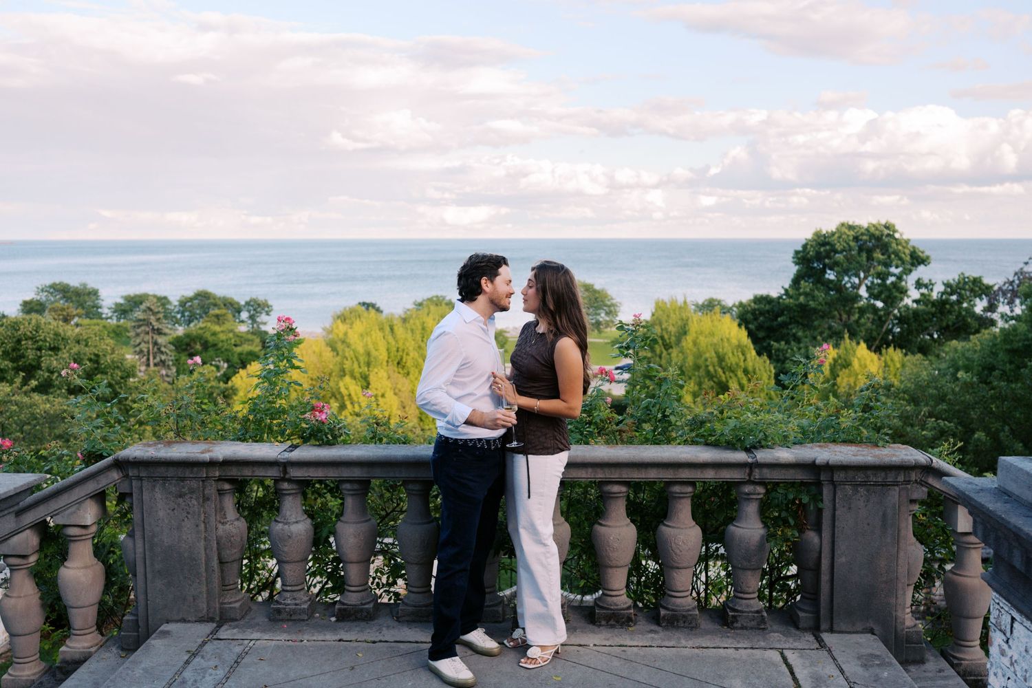 Couple sharing a romantic moment on a stone balcony overlooking lush gardens and the ocean at sunset.