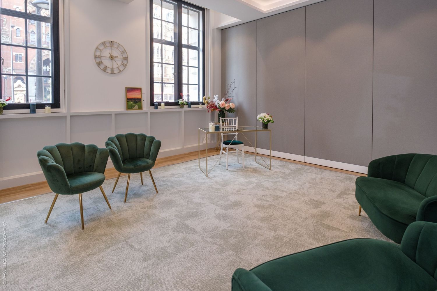 Elegant waiting room with green velvet chairs, gold accents, grey carpet, large windows, and a wall clock in a modern office.