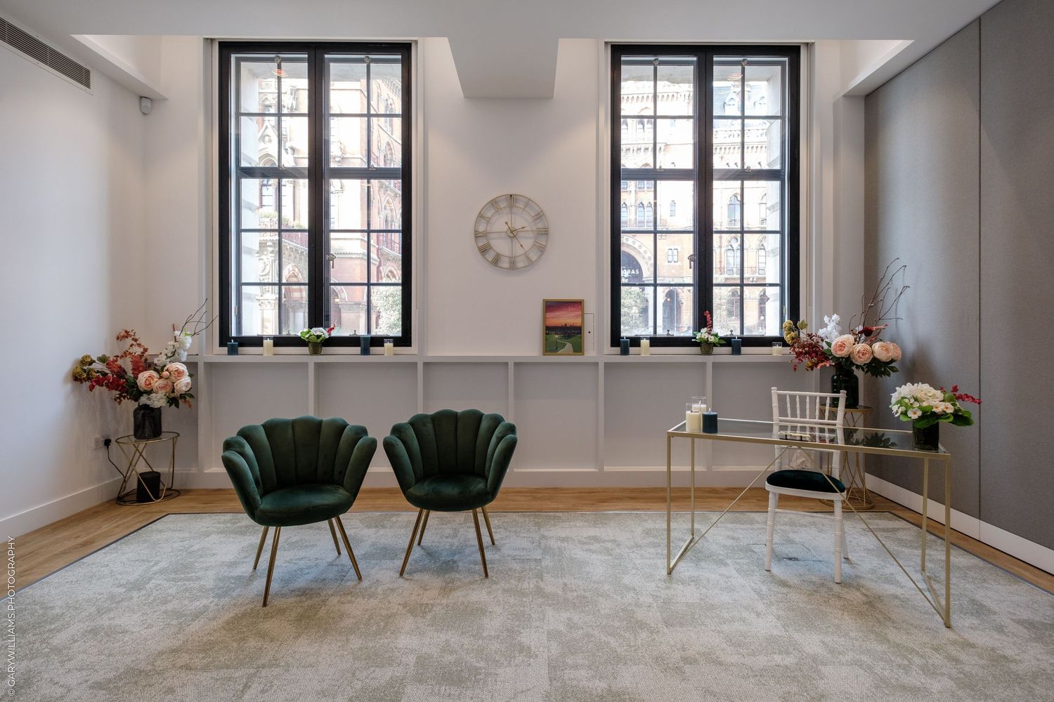 Elegant waiting room with two green velvet chairs, floral arrangements, and large black-framed windows in a modern interior.