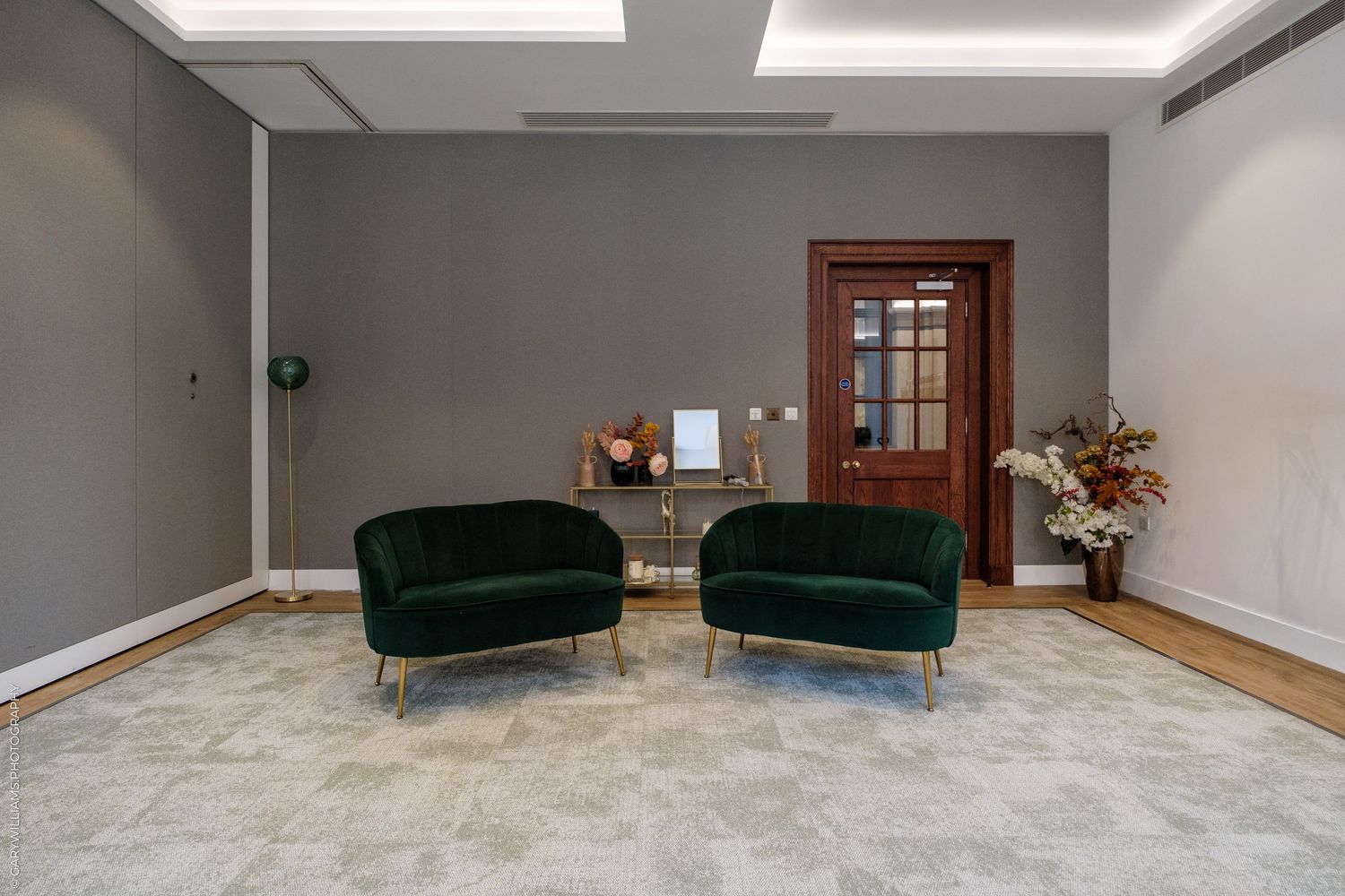 Elegant waiting area with two green velvet chairs, gold legs, gray walls, and a dark wood door.