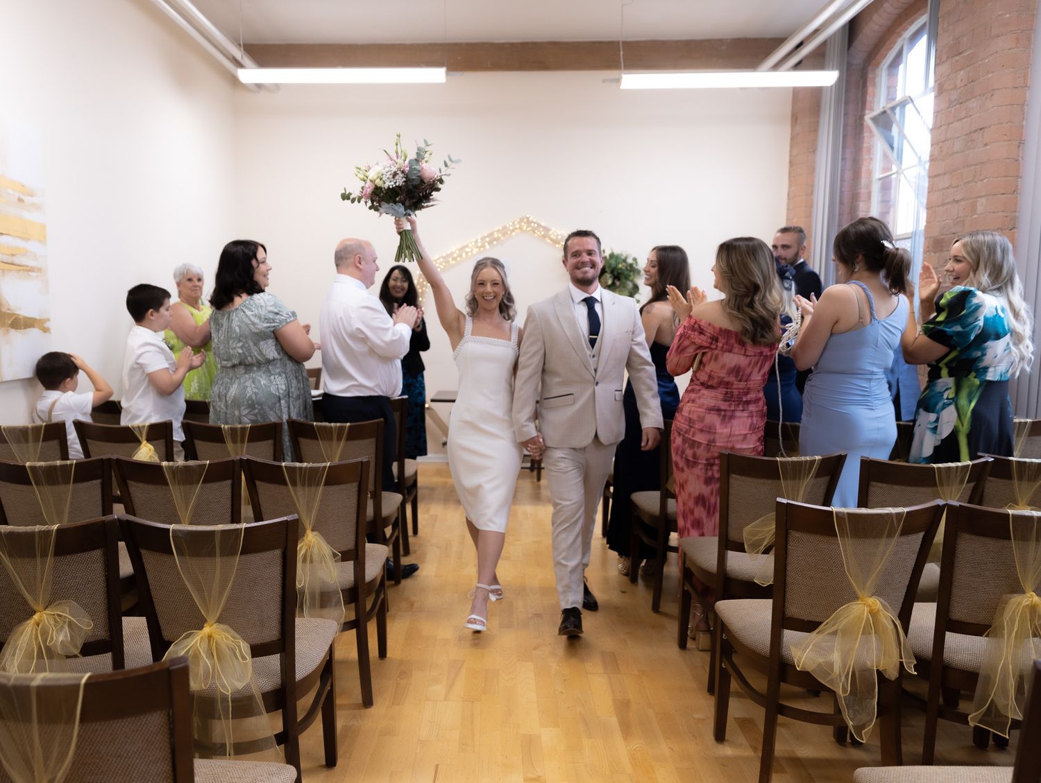 Newlywed couple walks down the aisle at indoor wedding ceremony, guests applauding, bride holding bouquet.