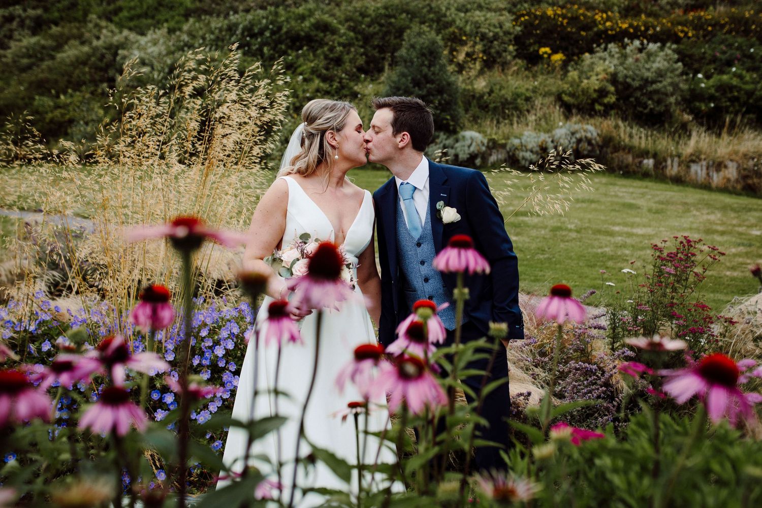 A bride and groom share a romantic kiss surrounded by pink coneflowers in a lush garden wedding setting.