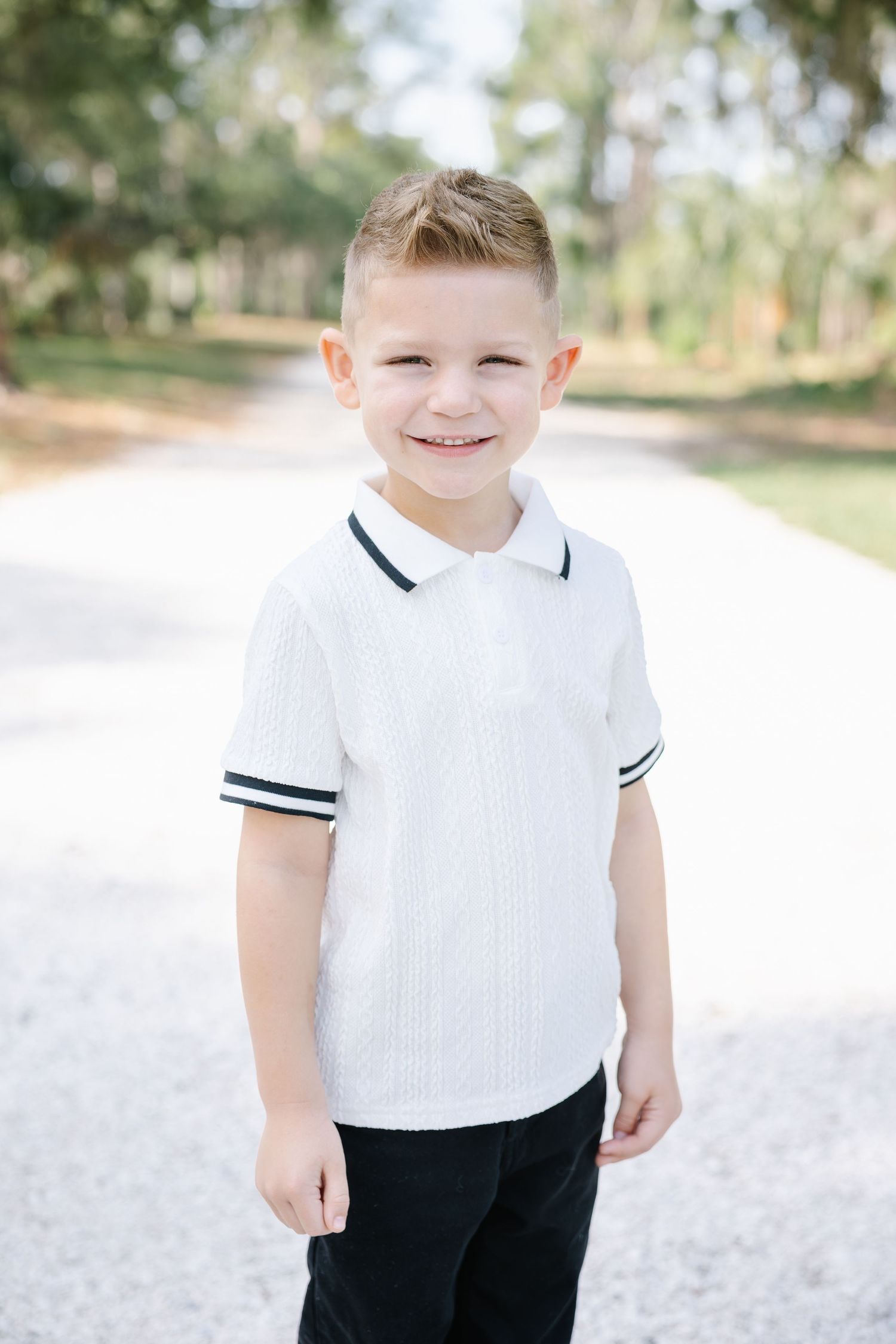 A cute young boy in a white polo shirt smiles on a sunny outdoor path.