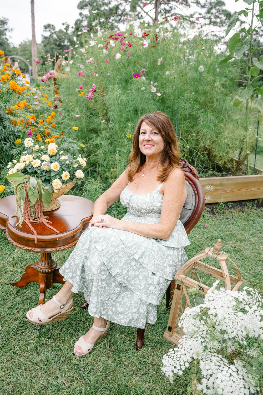 Smiling woman in floral sundress sits in vintage chair surrounded by colorful blooming garden flowers.