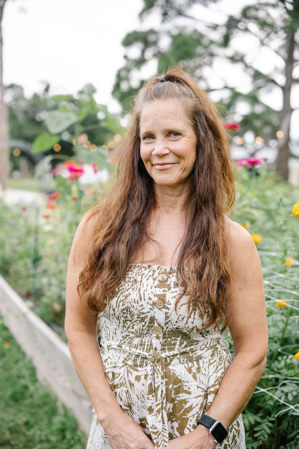 A smiling middle-aged woman with long brown hair poses in a floral sundress in a colorful garden.