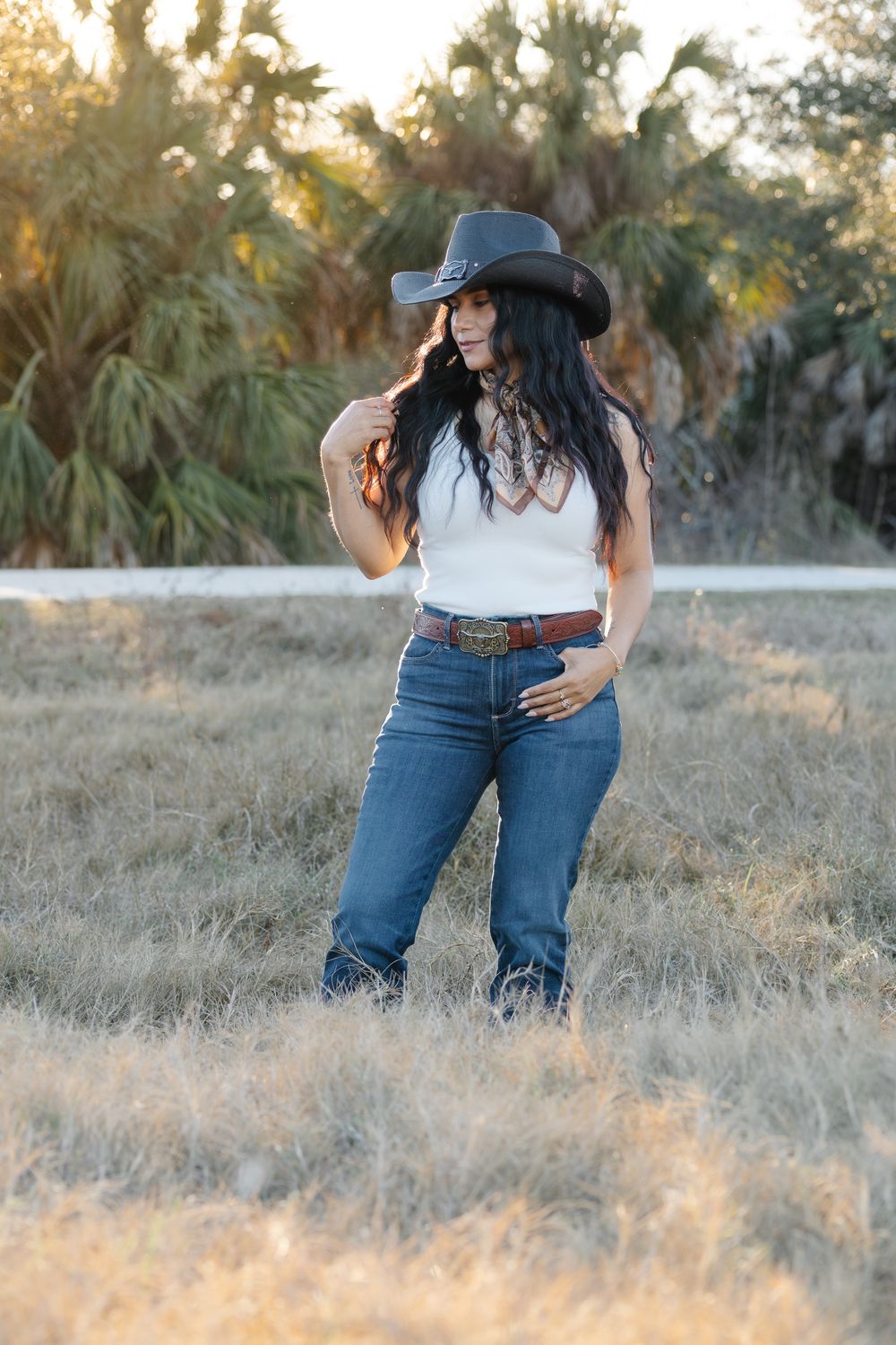 A woman in a cowboy hat, white top, and jeans poses in a dry field at sunset.