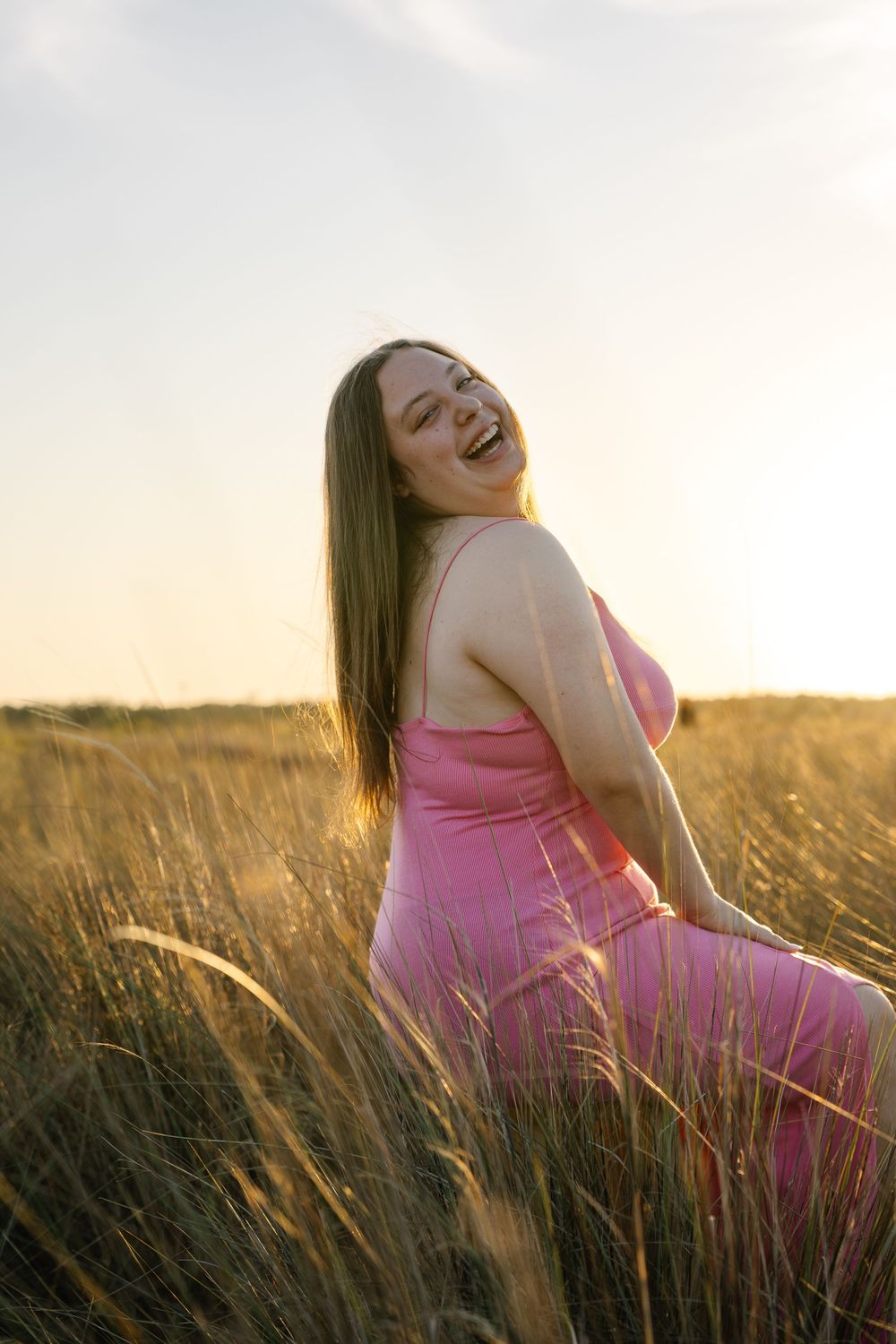 A joyful young woman in a pink dress laughs in a golden wheat field at sunset.