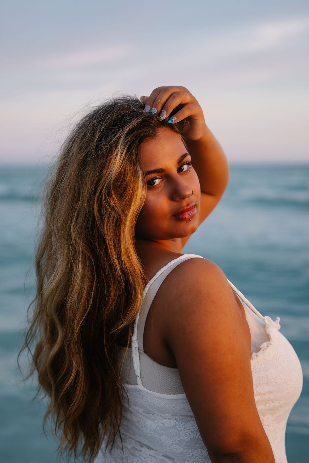 Young woman with long wavy hair poses at beach sunset in white top, hand in hair, ocean backdrop.