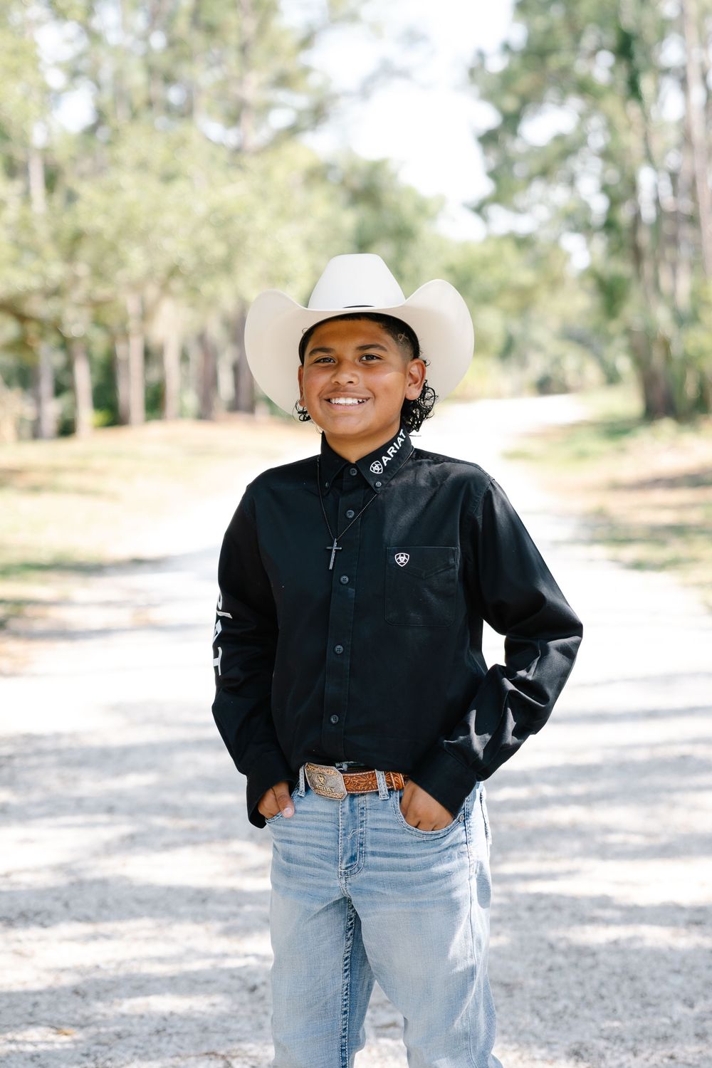 Smiling boy in white cowboy hat and black Ariat western shirt with jeans poses on a tree-lined dirt path.