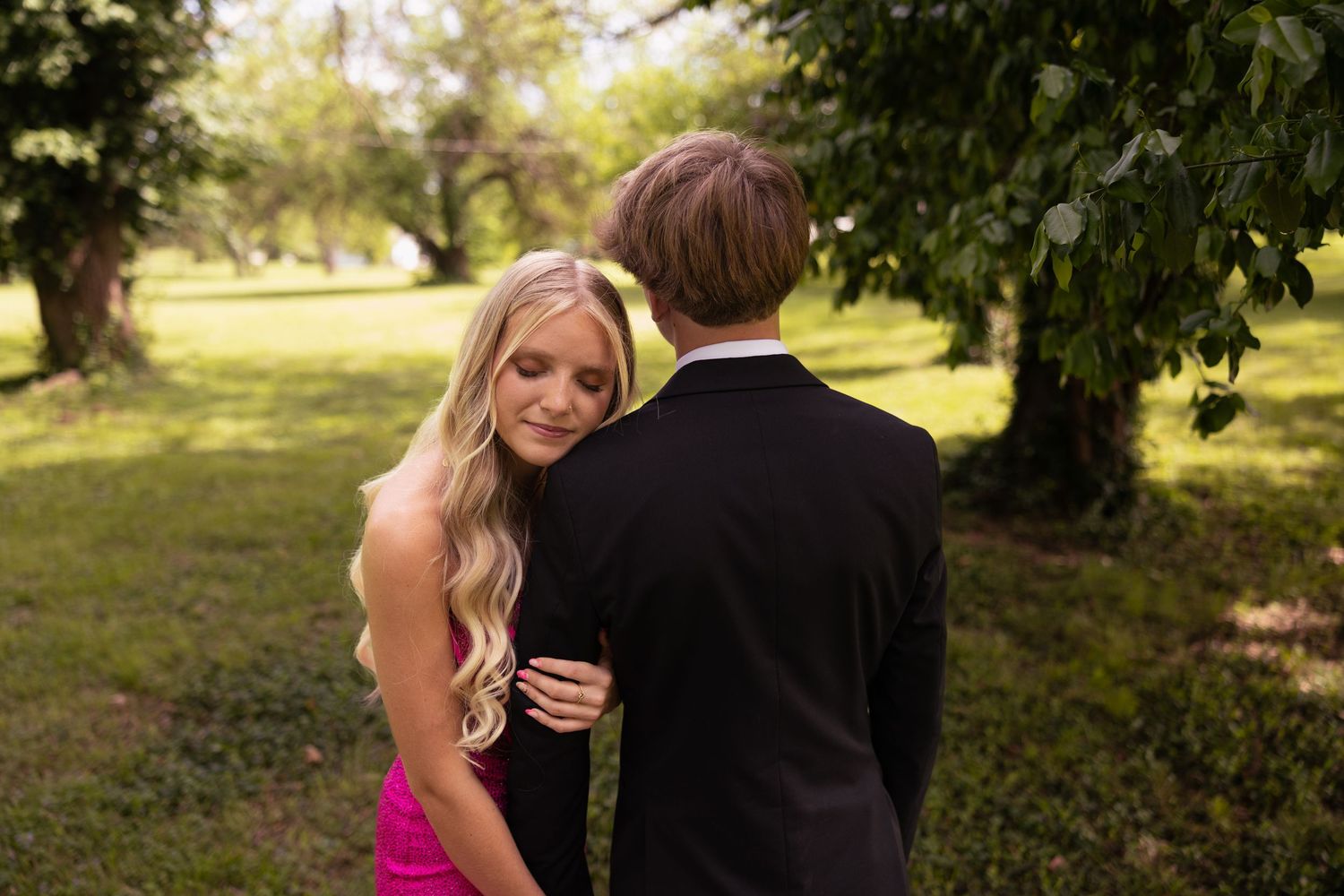 Couple in formal attire embracing outdoors, blonde woman in pink dress with man in black suit among green trees.