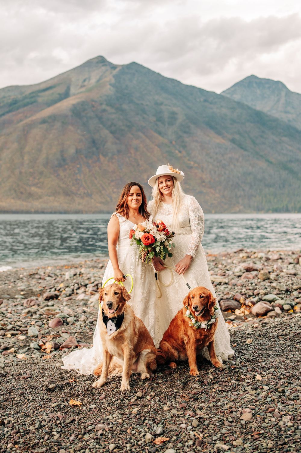 Two brides in white dresses pose with golden retrievers by a mountain lake on their wedding day.