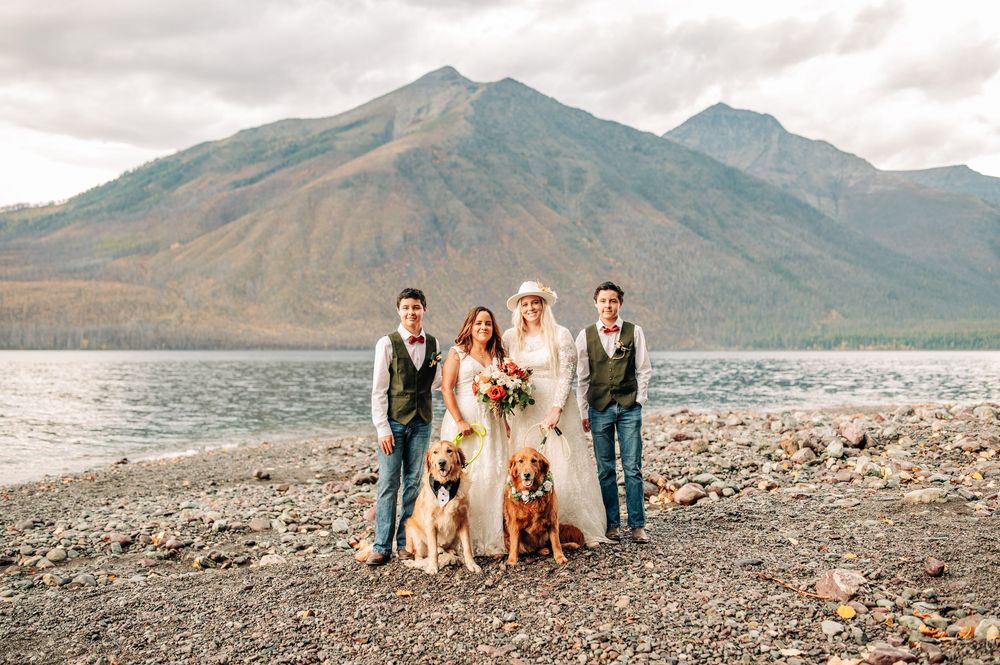 Wedding party with two golden retrievers poses on rocky lakeshore with mountain backdrop on cloudy day.