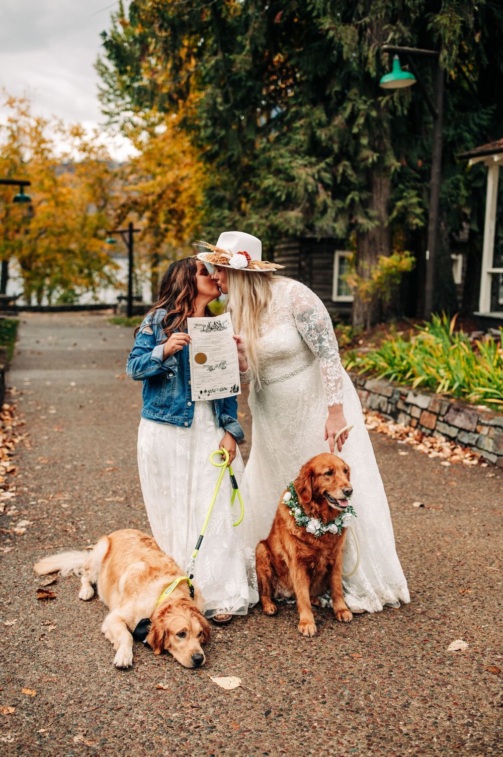 Two brides in white lace dresses kiss outdoors with their golden retrievers wearing floral collars on a fall wedding day.
