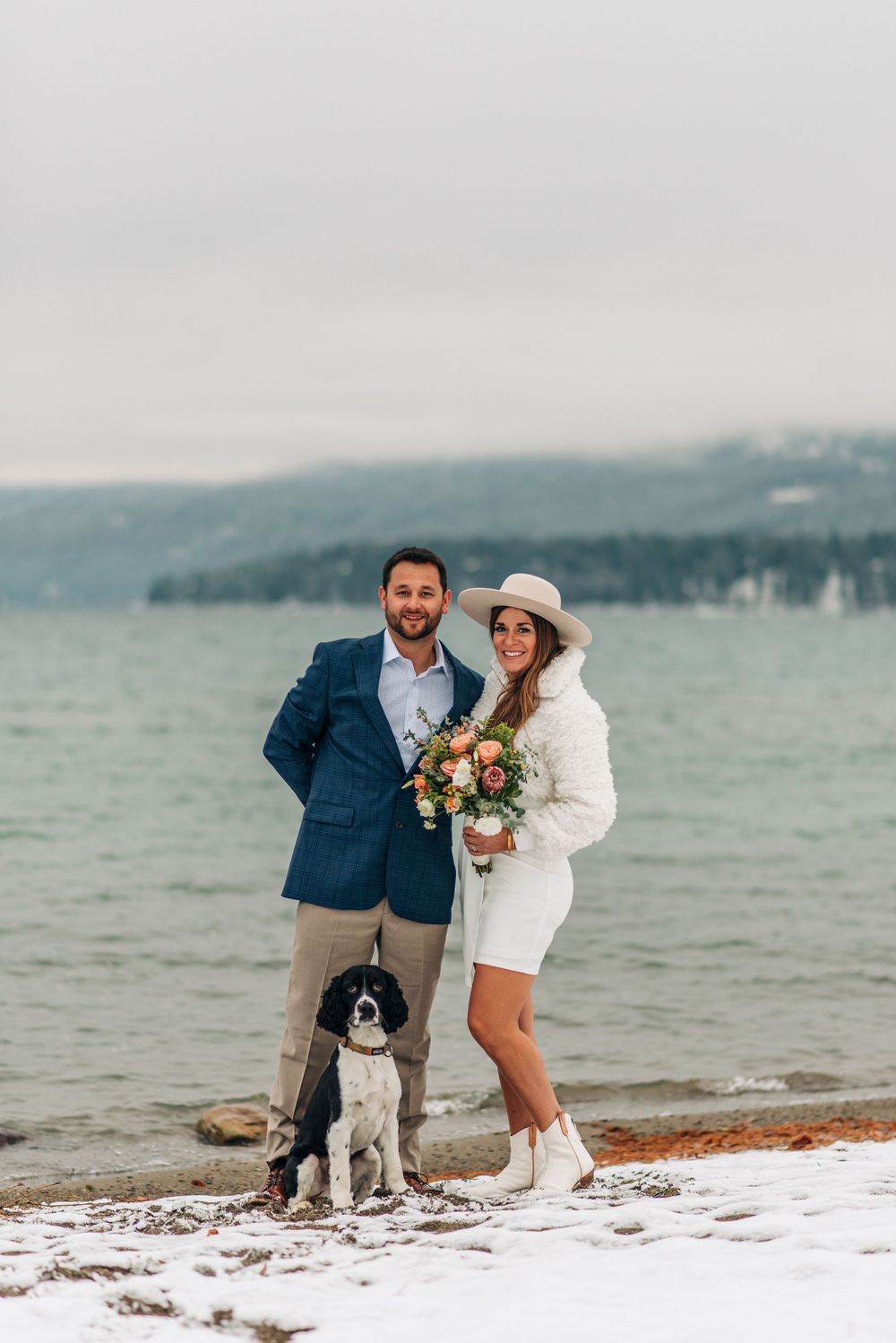 Couple in winter wedding attire with dog on snowy lakeshore, woman holding bouquet in white coat and hat.