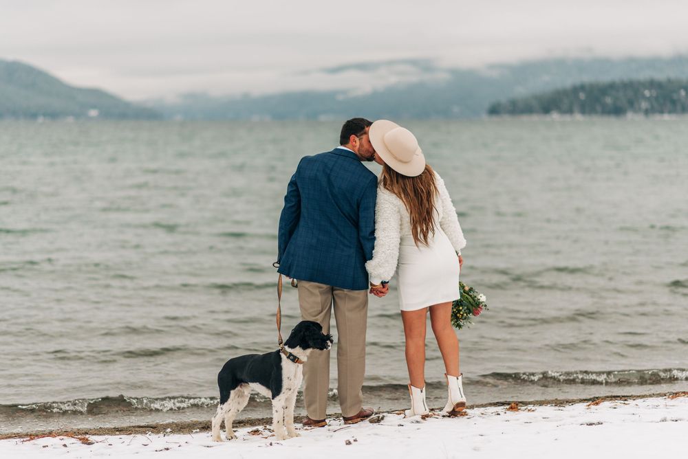 Couple in winter wedding attire standing on snowy beach with border collie dog, overlooking misty lake and mountains.
