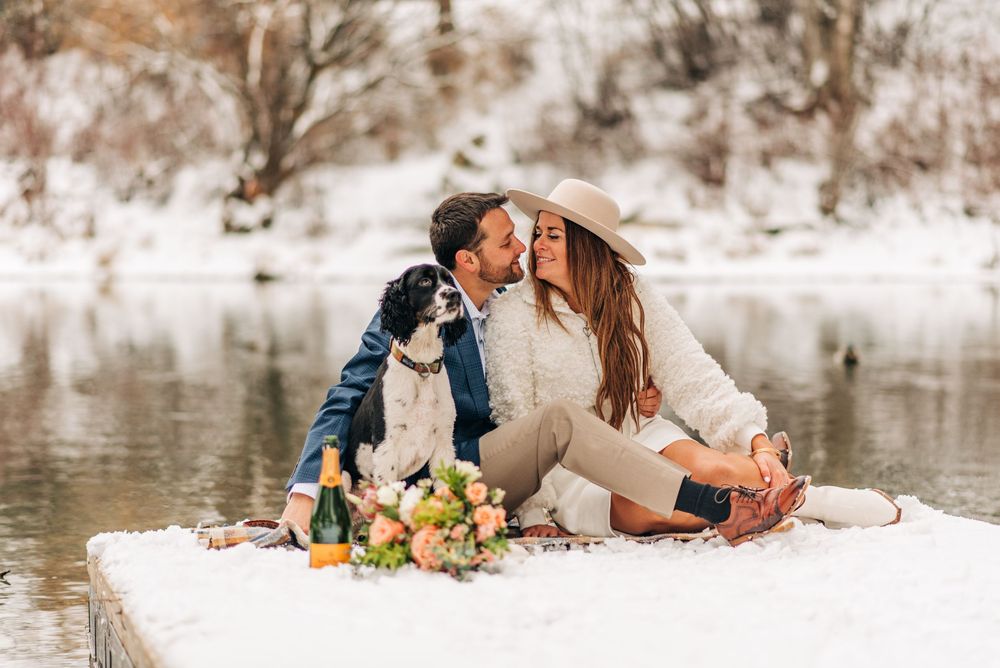 Couple kissing on snowy dock with dog and floral bouquet by a winter lake.