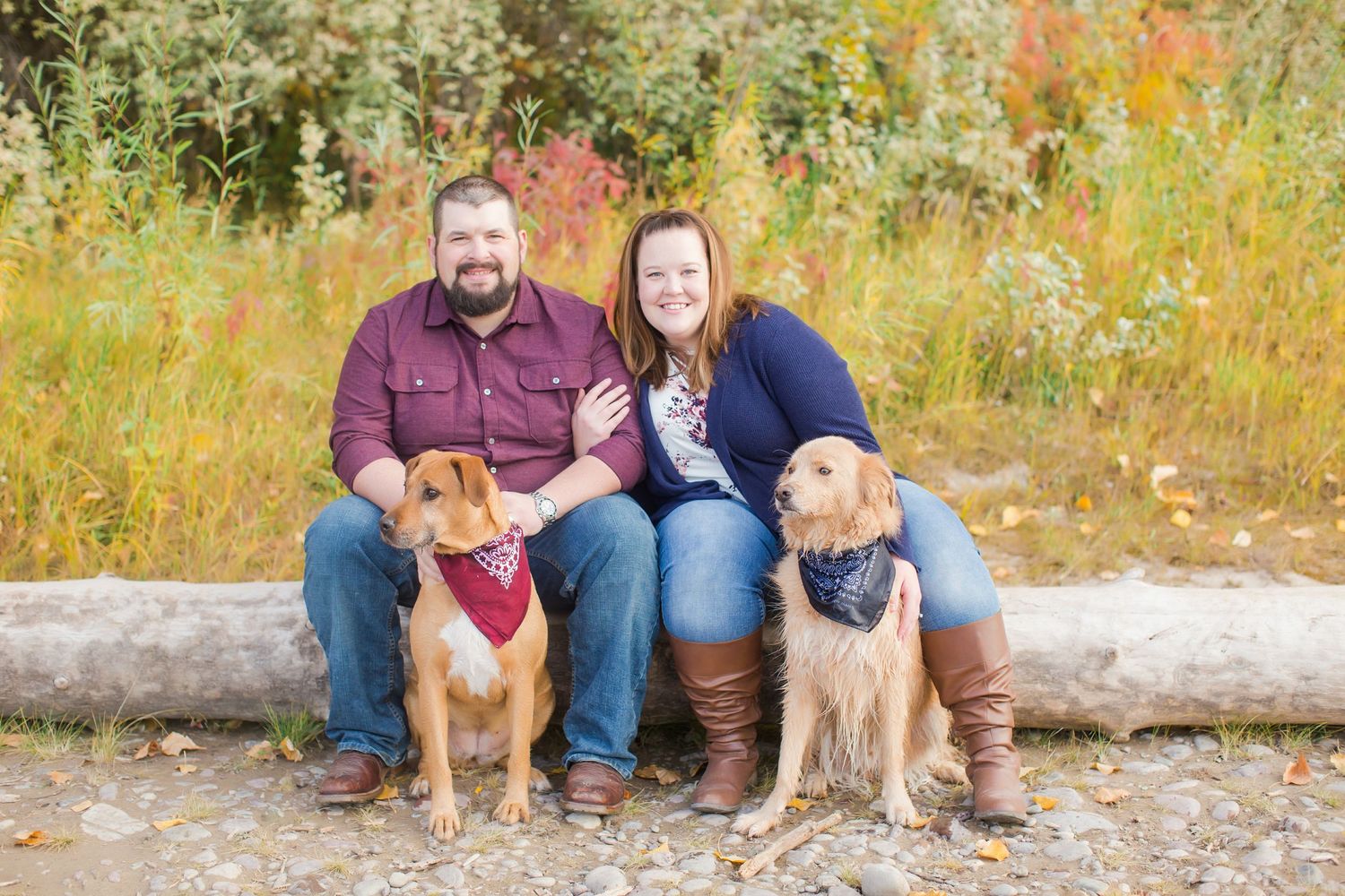 Smiling couple sitting outdoors with two dogs wearing bandanas, surrounded by colorful autumn foliage.