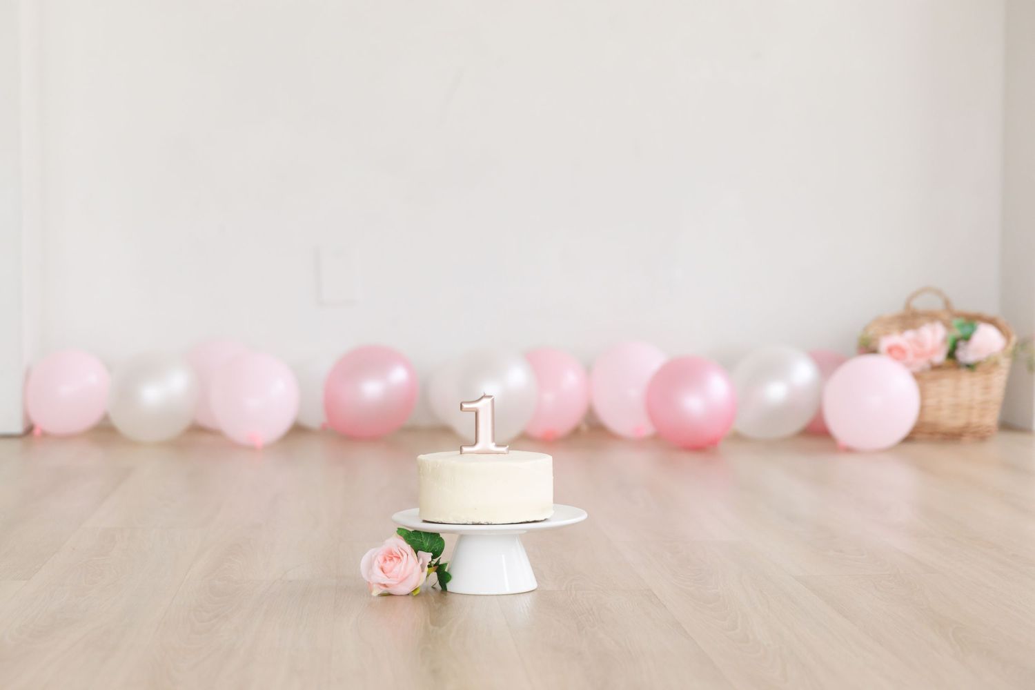 Baby girl in pink dress sits on hardwood floor between two wicker baskets of pink and white flowers.