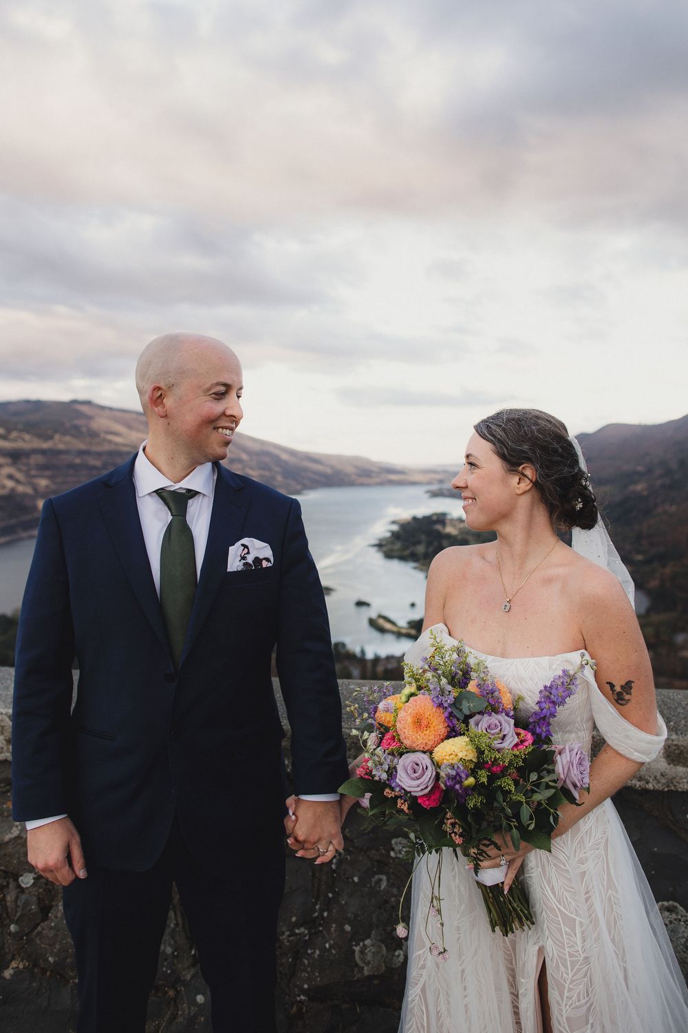 Bride and groom holding hands on a hilltop overlooking a lake, bride holding a colorful bouquet in a white gown.