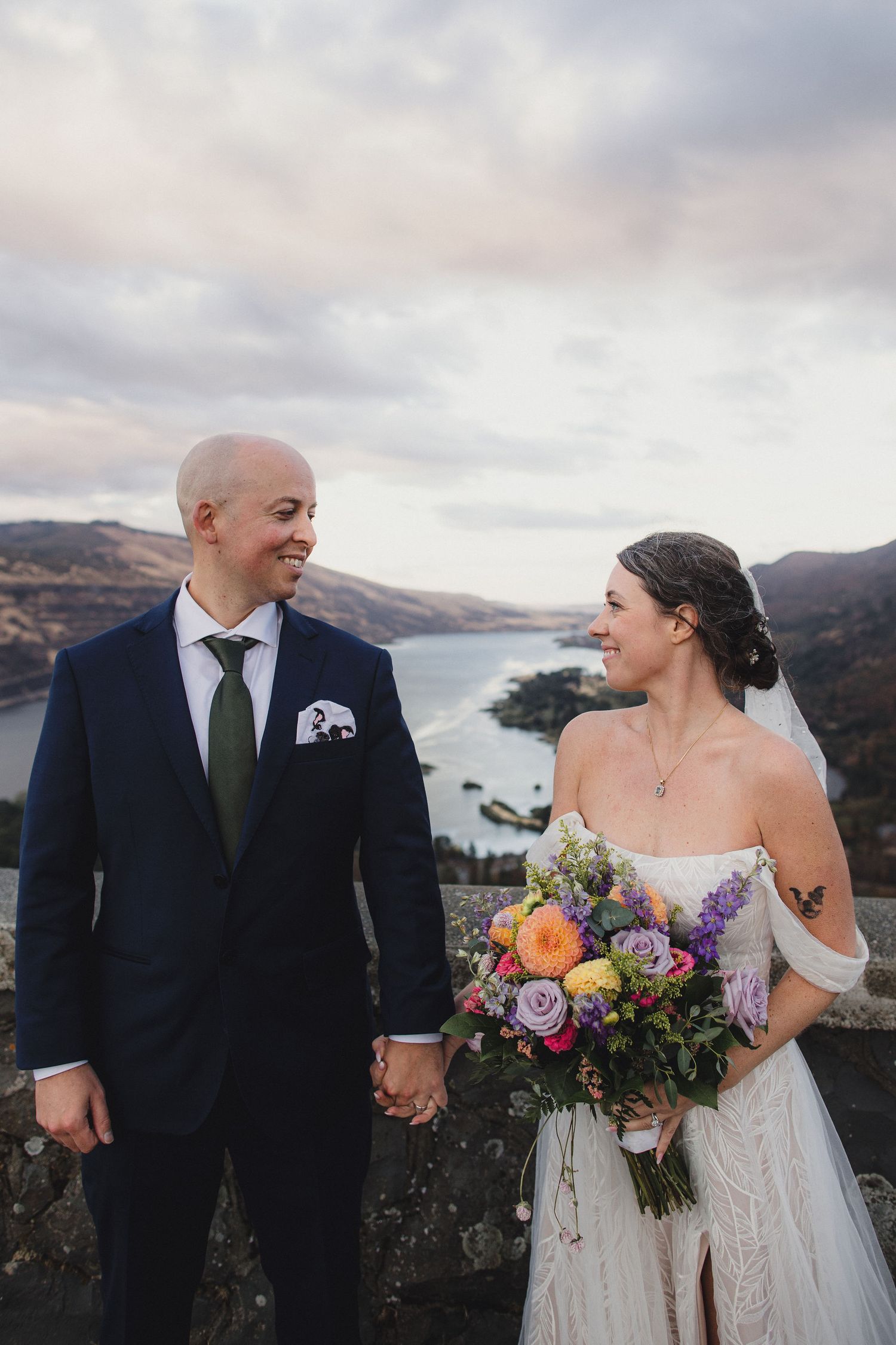 Bride and groom holding hands on a hilltop overlooking a lake, bride holding a colorful bouquet in a white gown.