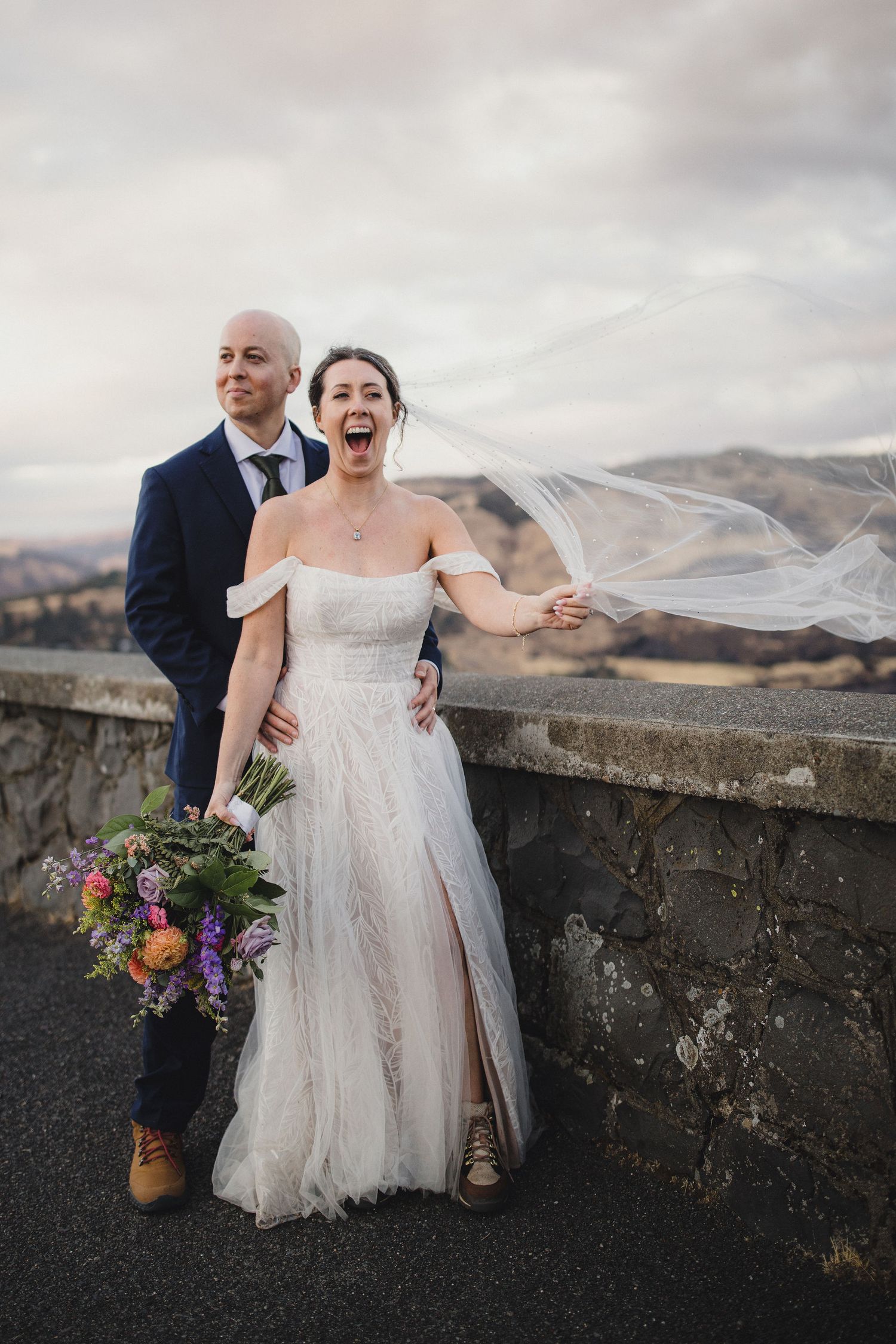Joyful bride in off-shoulder gown with flowing veil and groom in navy suit pose outdoors on their wedding day.